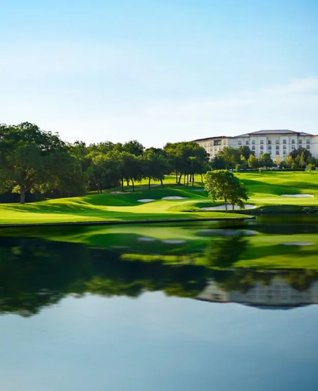A large building overlooks a lush green golf course with sand traps and a reflecting pond under a clear blue sky.