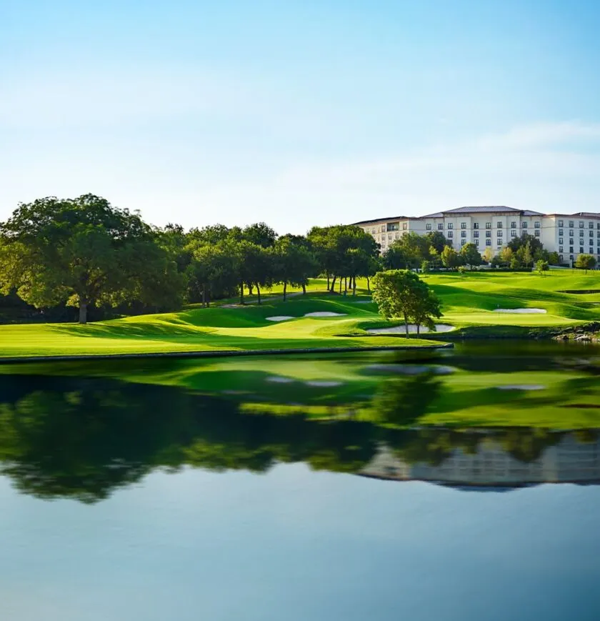 A large building overlooks a lush green golf course with sand traps and a reflecting pond under a clear blue sky.