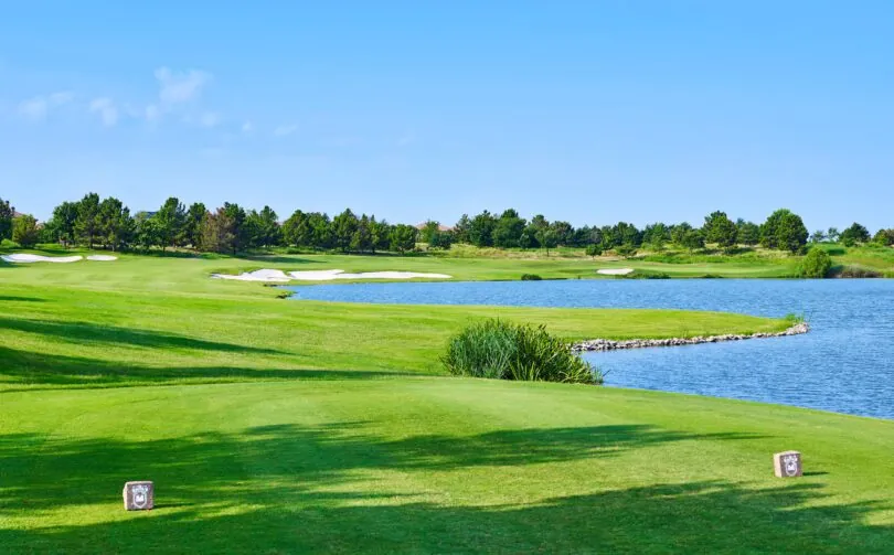 Bright golf course with green fairway, sand bunkers, and a blue water hazard under a clear sky, edged by trees in the distance.
