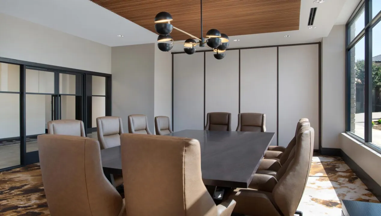 Modern conference room with a large dark table, ten brown chairs, geometric light fixture, and large windows letting in natural light.