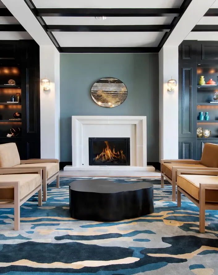 Modern lounge with tan chairs, geometric tables, a fireplace, built-in shelves, and abstract decor on blue-patterned carpet.