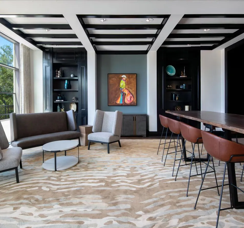 Modern lounge with gray chairs, a brown sofa, round tables, high stools at a wooden bar, built-in shelves, large windows, and colorful artwork.