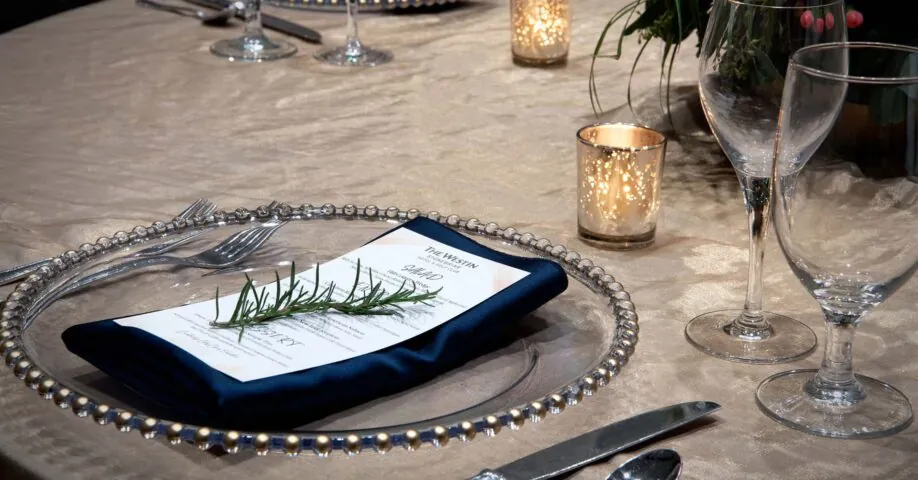 Elegant table setting with a beaded charger, menu, navy napkin, rosemary sprig, wine glasses, candles, silverware, and a floral centerpiece.