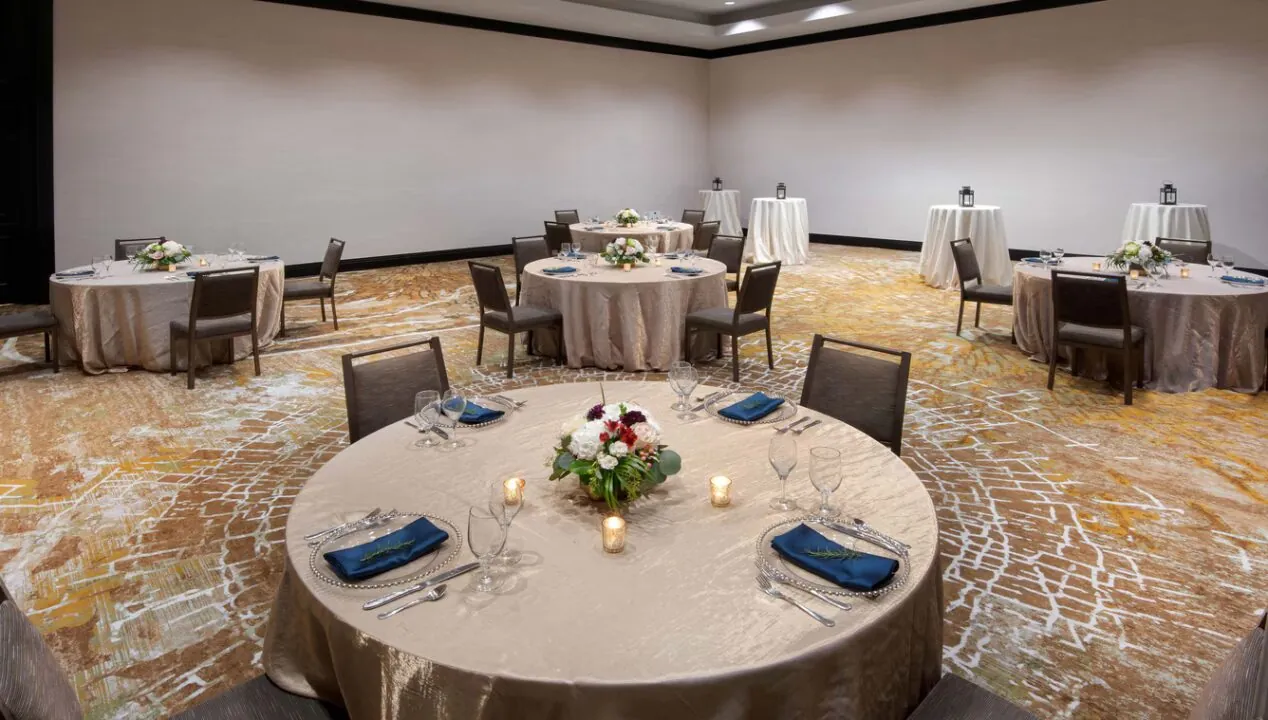 A banquet room with round tables covered in beige cloths, each set for four guests, spaced apart with floral centerpieces and candles.