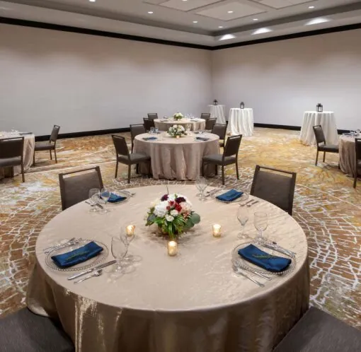 A banquet room with round tables covered in beige cloths, each set for four guests, spaced apart with floral centerpieces and candles.
