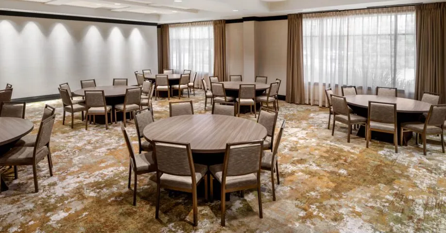 Spacious, well-lit conference room with round tables and chairs, large windows with curtains, and patterned carpet.