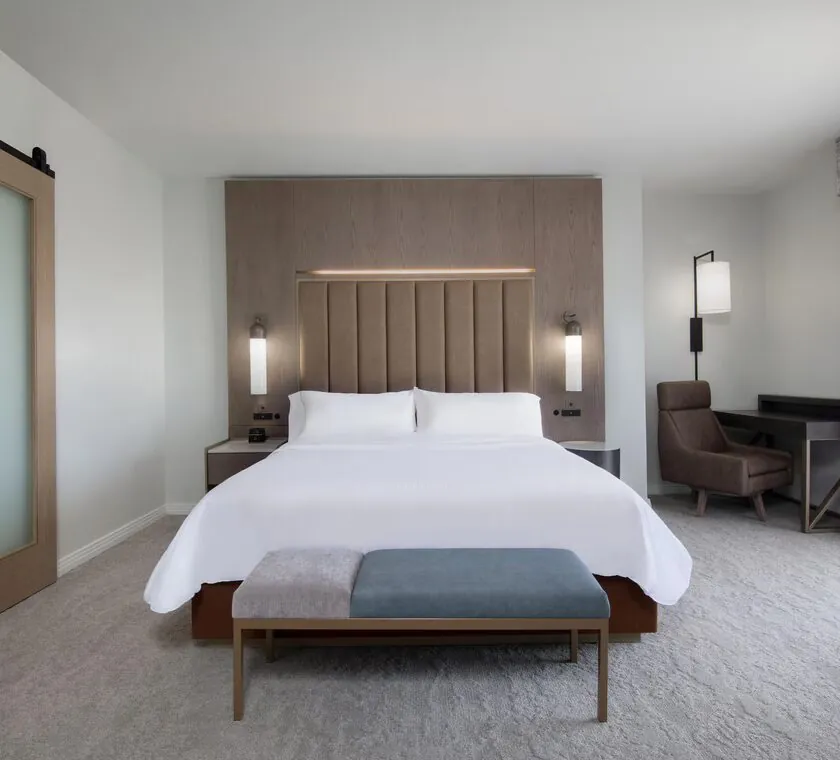 Modern hotel room with a king bed, padded headboard, bedside lights, small bench, desk, and large window with sheer curtains.