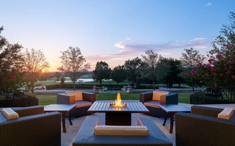 Wicker patio chairs surround a fire pit on a terrace, overlooking a golf course and trees at sunset.