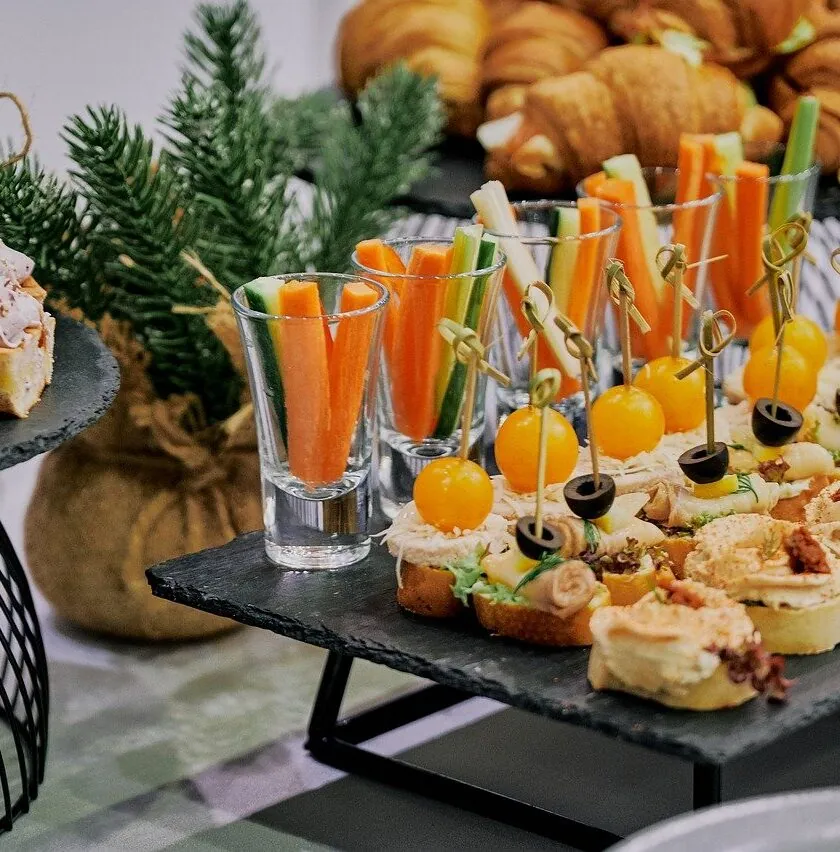 Assorted appetizers, including canapés, vegetable sticks in glasses, and croissants, arranged on serving trays with a small decorative pine tree.