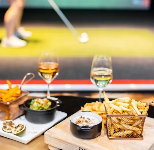 A table with fries, dip, snacks, and two glasses of wine, with a person playing indoor golf in the blurred background.