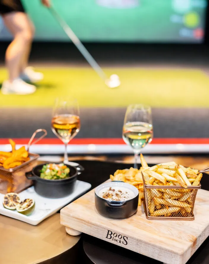 A table with fries, dip, snacks, and two glasses of wine, with a person playing indoor golf in the blurred background.