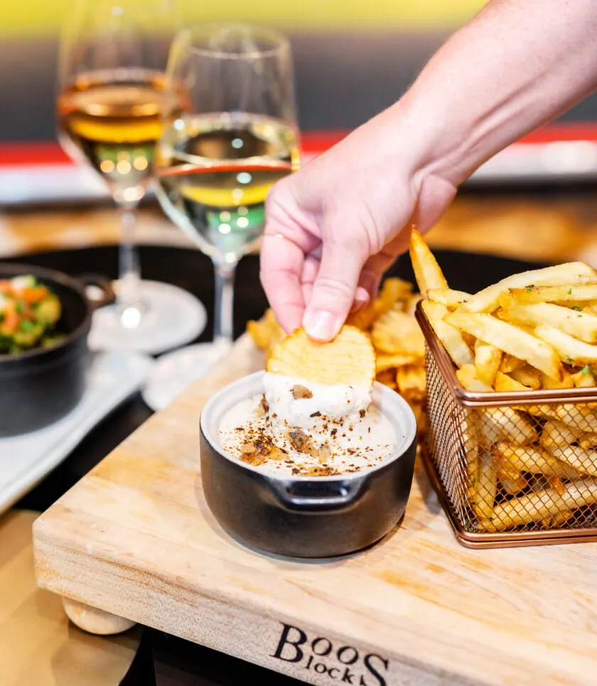 A hand dips a potato chip into a creamy dip beside fries, two glasses of wine, and a small bowl of food on a wooden board.