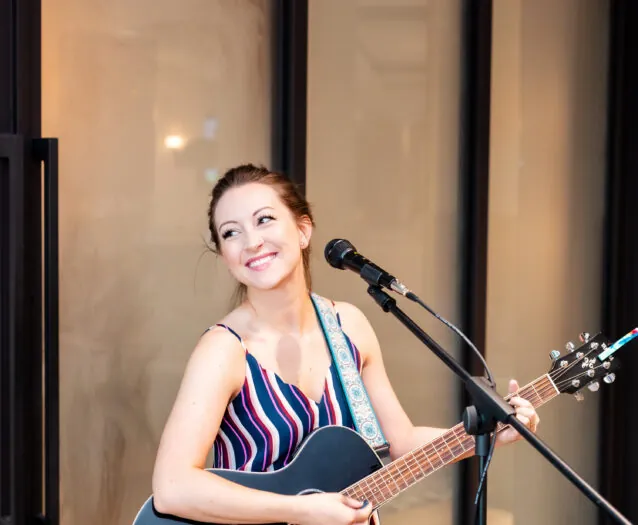 A woman smiles while playing a black acoustic guitar and singing into a microphone, standing indoors in front of large glass doors.